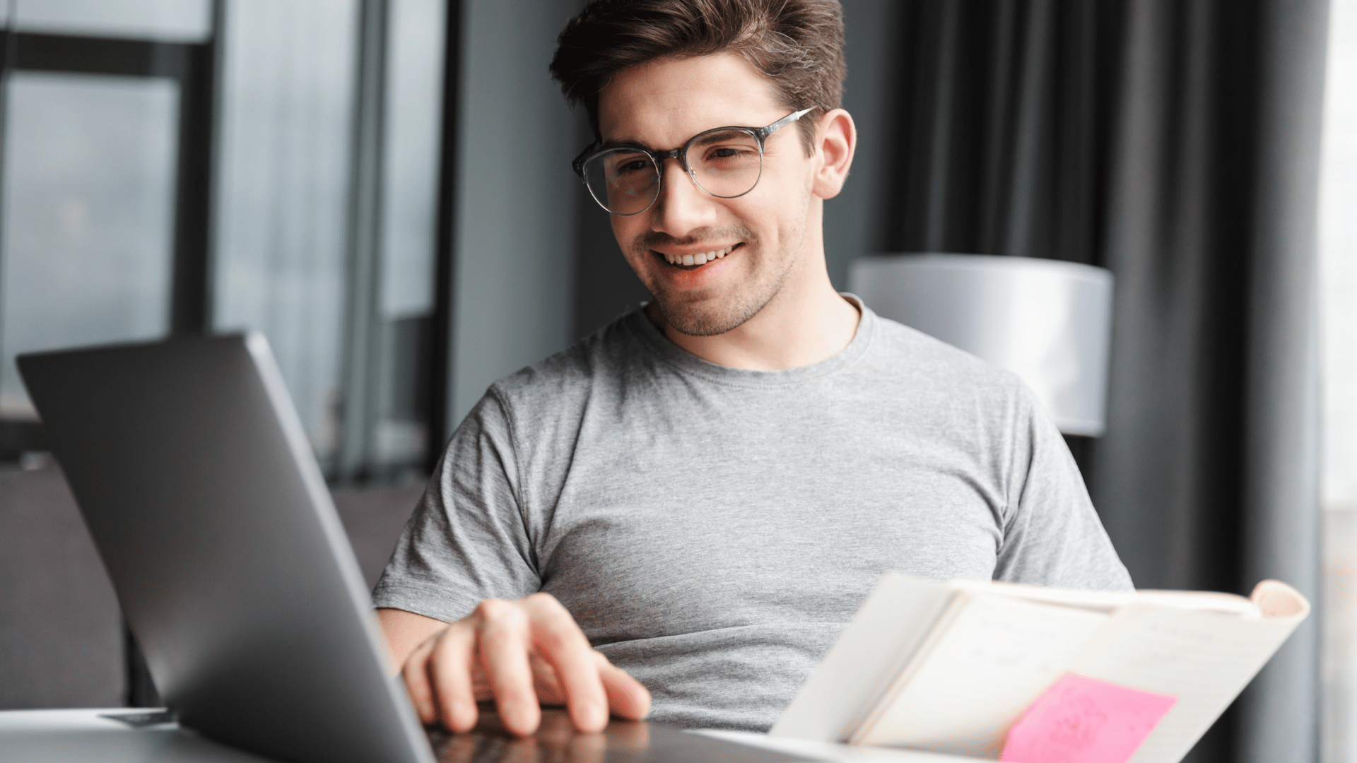 Homem jovem sorrindo enquanto trabalha em um notebook, usando camiseta cinza e óculos, segurando um caderno com anotações, em ambiente moderno e iluminado.