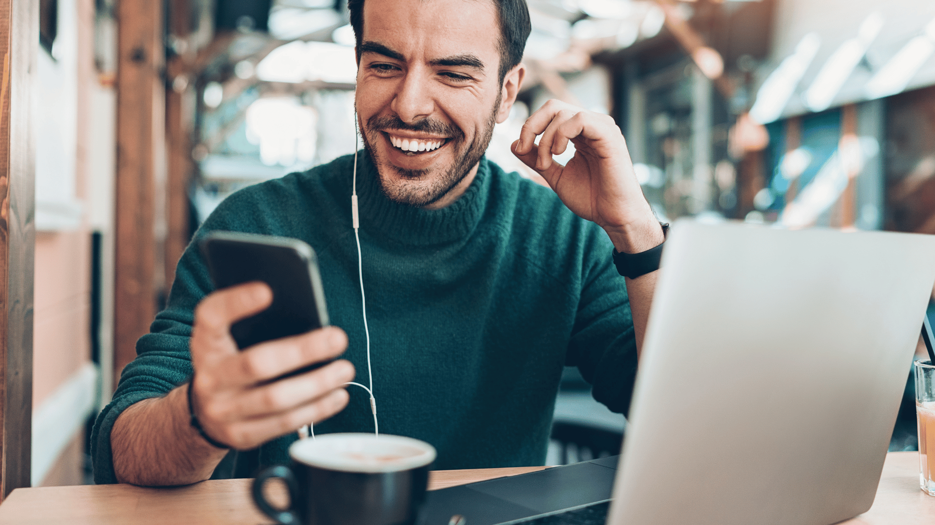 Homem sorrindo enquanto usa smartphone e notebook em cafeteria, conectado com fones de ouvido